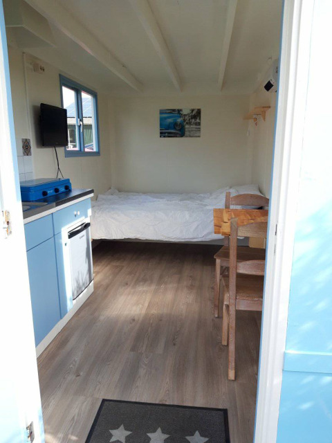 Vista interior de Pipowagen Snuitje en Camping De Gouwe Stek, cama, cocina pequeña y muebles de madera.