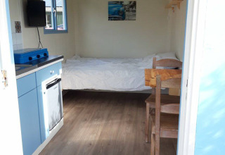 Vista interior de Pipowagen Snuitje en Camping De Gouwe Stek, cama, cocina pequeña y muebles de madera.