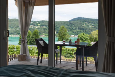 Vista da una tenda glamping in un parco vacanze con tavolo, sedie e paesaggio di lago e colline verdi.