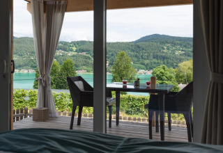 Zicht vanuit een glampingtent op een vakantiepark met tafel, stoelen en een meer met groene heuvels.