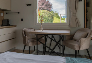 Moderne glamping accommodatie met twee zachte stoelen, een tafel en een raam met zicht op groene natuur.
