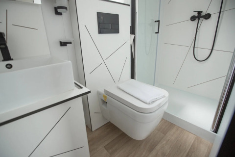 Baño moderno con inodoro suspendido, ducha, lavabo y suelo de madera clara en alojamiento glamping.