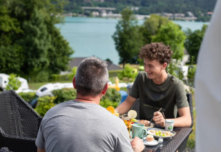 To personer nyder morgenmad udendørs med udsigt over en sø og glamping-vogne i en feriepark.