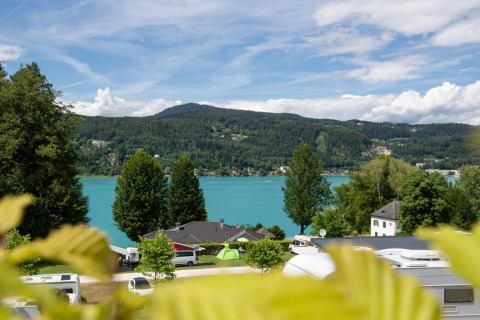 Vista de un lago y montañas desde un parque vacacional con tiendas glamping y caravanas bajo cielo azul.