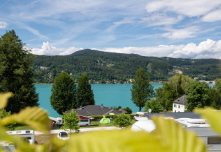 Blick auf einen See und Berge von einem Ferienpark mit Glamping und Wohnwagen unter blauem Himmel.