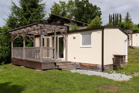 Glampingverblijf met houten terras en veranda in een groene vakantiewijk omgeven door bomen.