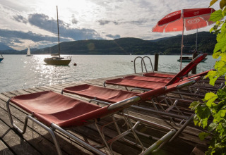 Ligstoelen en een rode parasol op een houten ponton aan het meer, met zeilbootjes en heuvels in de verte.