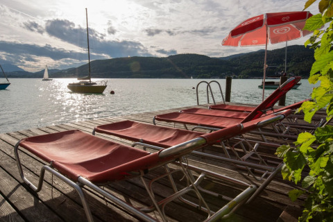 Sun loungers and a red umbrella on a wooden dock by the lake, with sailboats and hills in the background.