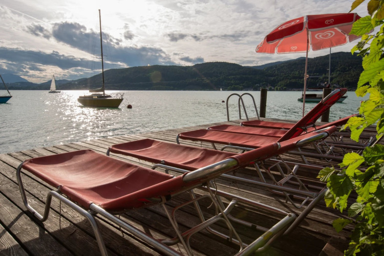 Sun loungers and a red umbrella on a wooden dock by the lake, with sailboats and hills in the background.