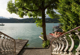Blick von einer Treppe auf einen See mit Baum, Holzdeck und Person auf Liege im Glamping-Resort.