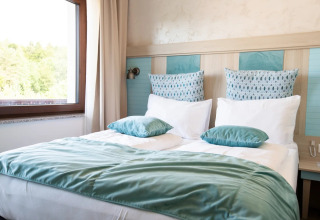 Lit douillet avec oreillers et couverture bleus près de la fenêtre, logement glamping dans un parc de vacances.