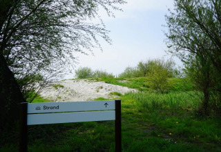 Pad tussen groene bomen naar zandstrand in vakantiepark met glamping, strandbord vooraan zichtbaar.