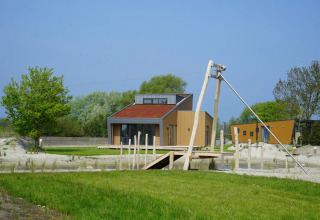 Et moderne glampinghus med rød tag ved en ferielejr, omgivet af grønne træer og åbne områder under blå himmel.