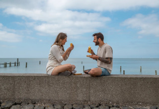 Un couple partage un repas près de la mer dans un parc de vacances avec glamping sous un ciel bleu.