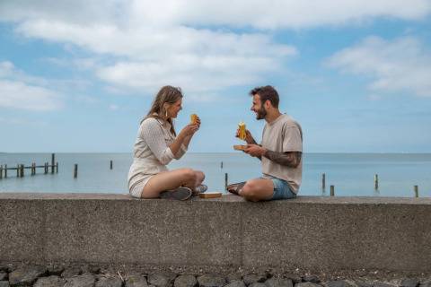 Un couple partage un repas près de la mer dans un parc de vacances avec glamping sous un ciel bleu.