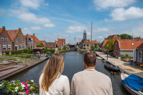 Pareja observa un canal en parque vacacional con glamping, casas coloridas y barcos en un día soleado.