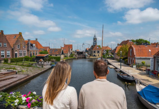 Pareja observa un canal en parque vacacional con glamping, casas coloridas y barcos en un día soleado.