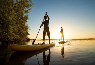 Two people paddleboarding on a calm lake at sunset near trees, enjoying nature at a glamping holiday park.