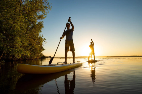 To personer står på paddleboards på en sø ved solnedgang nær træer, afslappende ferie i naturen.