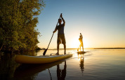 Two people paddleboarding on a calm lake at sunset near trees, enjoying nature at a glamping holiday park.