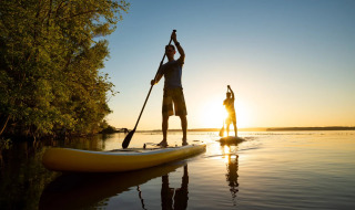 Dos personas hacen paddle surf al atardecer en un lago tranquilo junto a árboles, disfrutando del glamping.