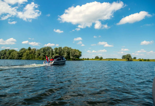 Un bateau à moteur navigue sur un lac sous un ciel bleu nuageux, entouré d’arbres dans un parc de vacances.