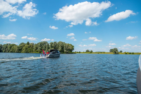 Una barca a motore naviga su un lago sotto un cielo azzurro con nuvole, circondata da alberi in un villaggio vacanze.
