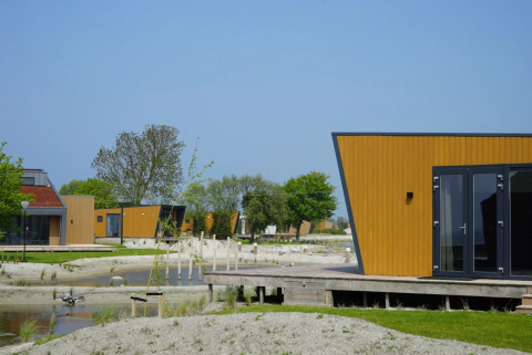Cabañas glamping modernas en un parque vacacional, rodeadas de arena, agua y vegetación bajo cielo azul.