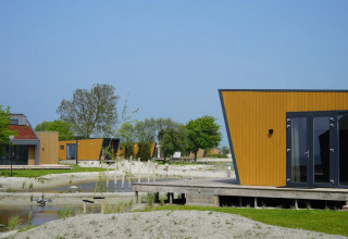 Cabanes de glamping modernes dans un parc de vacances avec sable, eau et espaces verts sous ciel bleu.
