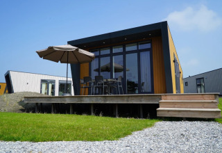 Moderne glampinghut in vakantiepark met verhoogd terras, tuinset en parasol op gazon onder blauwe lucht.