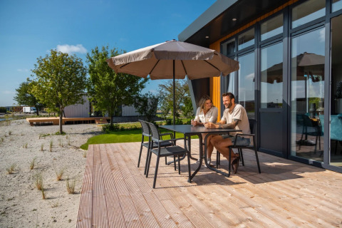 Paar sitzt unter Sonnenschirm auf Holzterrasse eines modernen Glamping-Ferienhauses im Park.