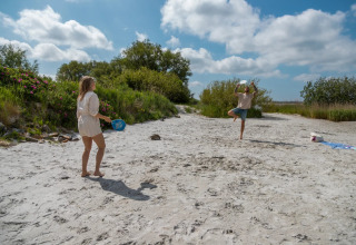 Twee personen spelen tennis op het strand bij een vakantiepark met glamping accommodaties.