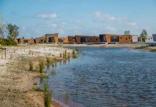 Cabanes de glamping modernes près d’un lac dans un parc de vacances, entourées de sable et de verdure.