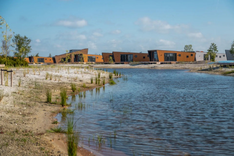 Modern glamping cabins by a lake in a holiday park, surrounded by sand, grass, and young trees.