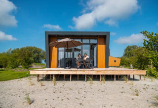 Moderne glamping lodge met houten terras en parasol, twee personen genieten bij een vakantiepark.