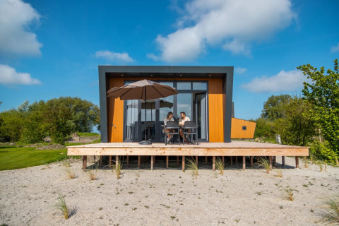 Moderne glamping lodge met houten terras en parasol, twee personen genieten bij een vakantiepark.