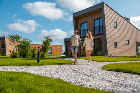 Par går hånd i hånd på en sti foran moderne glamping-lejeboliger i feriepark på en solrig dag.
