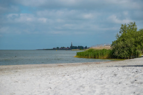 Photo of a calm beach with reeds and trees, a glamping holiday park, and a lighthouse in the background.