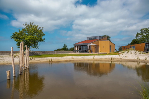 Photo of a holiday park with glamping lodges, a sandy pond, wooden houses, trees, and a blue sky.