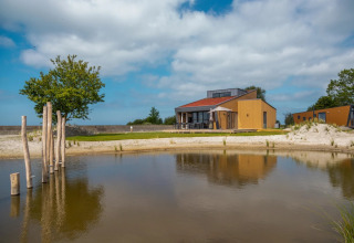 Photo of a holiday park with glamping lodges, a sandy pond, wooden houses, trees, and a blue sky.