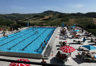 Piscina all'aperto con bagnanti e ombrelloni circondata da colline panoramiche in Lombardia, Italia.