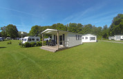 Lyst lodgehus med terrasse på grøn campingplads hos Camping Roland, Holland, omgivet af træer og natur.