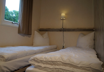 To enkeltsenge på et lille værelse med hvidt sengetøj, natlampe og vindue på Lodge at Camping Roland.
