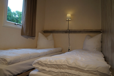 Zwei Einzelbetten mit weißer Bettwäsche in einem kompakten Zimmer, Lampe und Fenster im Camping Roland.