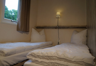 Zwei Einzelbetten mit weißer Bettwäsche in einem kompakten Zimmer, Lampe und Fenster im Camping Roland.