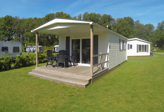 Modern lodge with covered wooden deck, outdoor table and chairs, set on a green lawn with trees nearby.