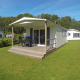 Moderne lodge med overdækket træterrasse, havemøbler og græsplæne, omgivet af natur og campingvogne.