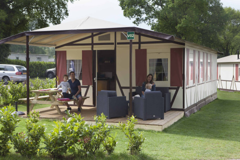 Famiglia rilassata fuori dal lodge Vakwerktent al Camping Roland, circondata dal verde nei Paesi Bassi.