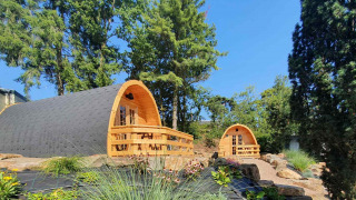 Dos pequeñas casas de madera llamadas Pods en un jardín verde con árboles altos y plantas diversas.