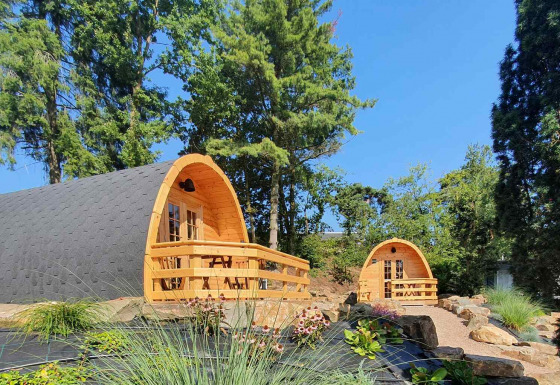 Dos pequeñas casas de madera llamadas Pods en un jardín verde con árboles altos y plantas diversas.